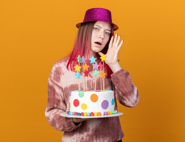 Suspicious young beautiful girl wearing party hat holding cake whispers isolated on orange wall