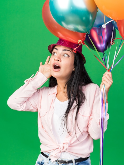 Подозрительная молодая красивая женщина в партийной шляпе держит воздушные шары, показывая жест слушания, изолированные на зеленой стене