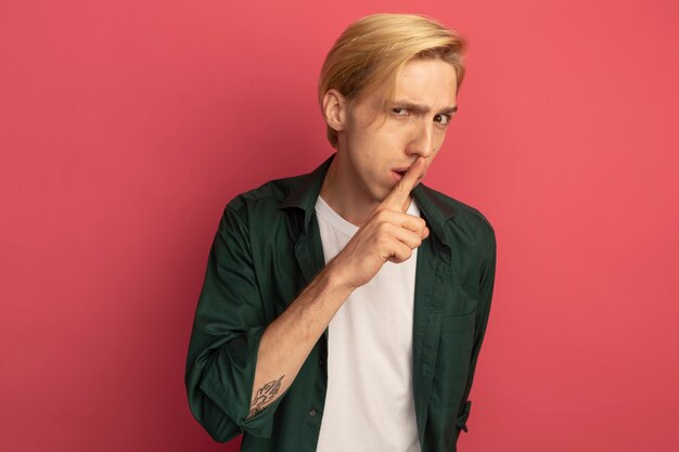 Suspicious young blonde guy wearing green t-shirt showing silence gesture