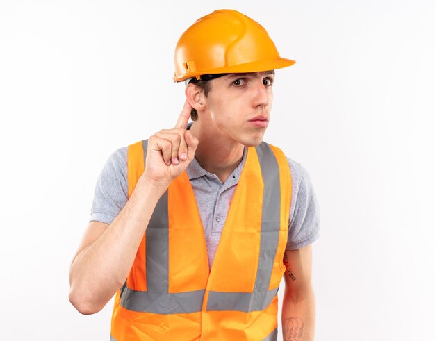 Suspicious young builder man in uniform showing listen gesture isolated on white wall