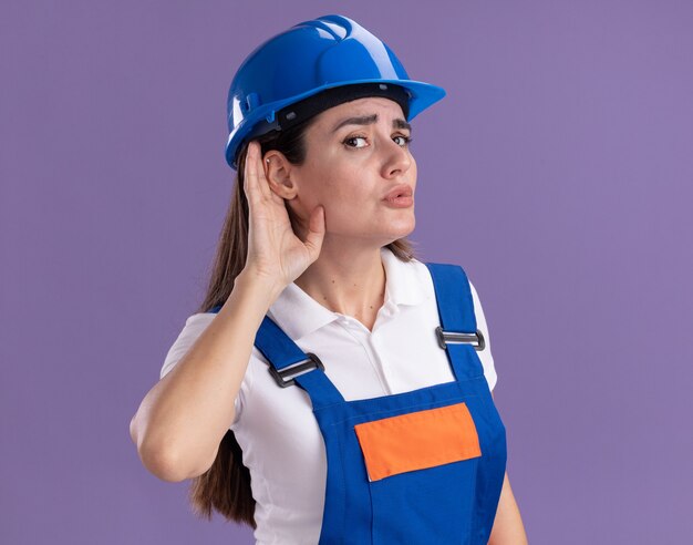 Подозрительная молодая женщина-строитель в униформе, показывающая жест слушания изолирована на фиолетовой стене