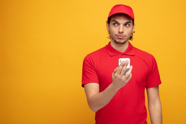 suspicious young delivery man wearing uniform and cap holding mobile phone looking at side isolated on yellow background with copy space