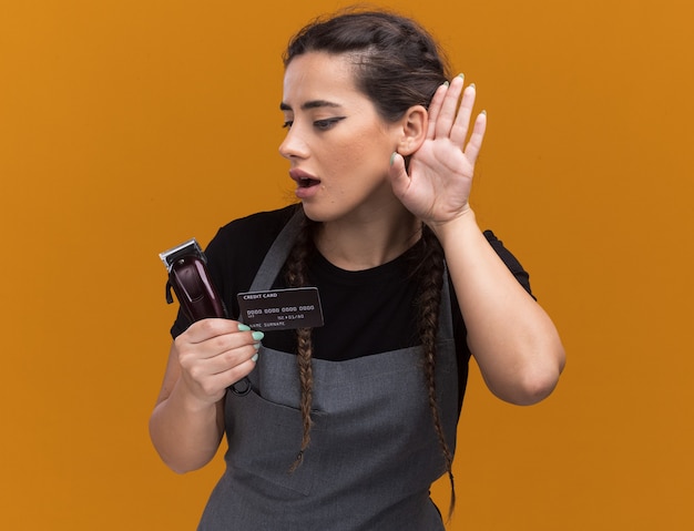 Giovane barbiere femminile sospettoso in uniforme che tiene la carta di credito e tagliacapelli che mostrano ascolta gesto isolato sulla parete arancione