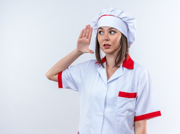 Suspicious young female cook wearing chef uniform showing listen gesture isolated on white background