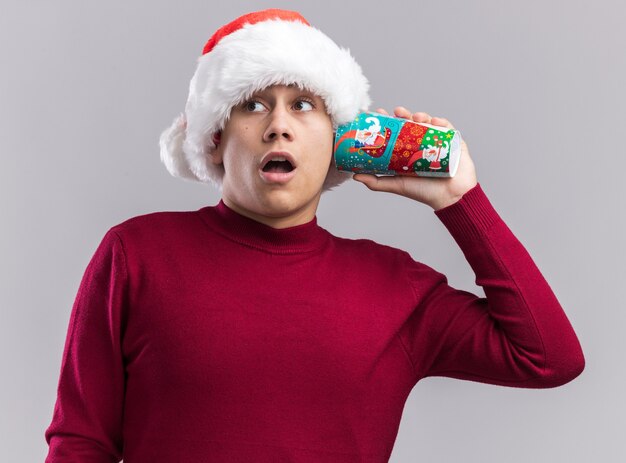 Suspicious young guy wearing christmas hat holding christmas cup on ear isolated on white wall