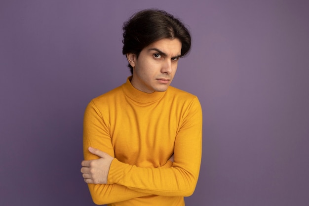 Suspicious young handsome guy wearing yellow turtleneck sweater crossing hands isolated on purple wall