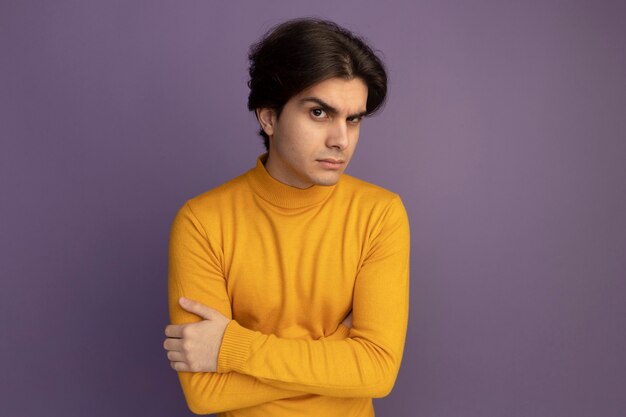 Suspicious young handsome guy wearing yellow turtleneck sweater crossing hands isolated on purple wall