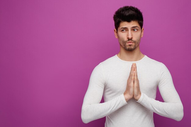 Suspicious young handsome man doing namaste gesture looking at side on purple background with copy space