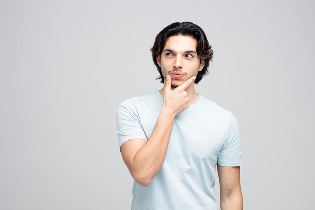 Suspicious young handsome man keeping hand on chin looking at side isolated on white background with copy space