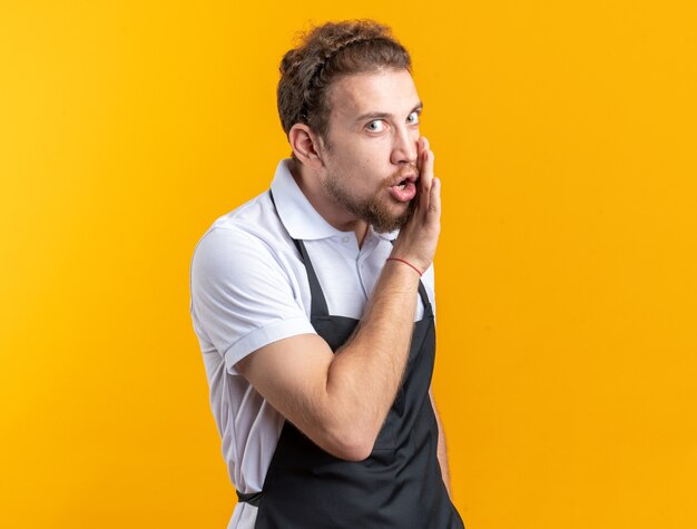 Suspicious young male barber wearing uniform whispers isolated on yellow background
