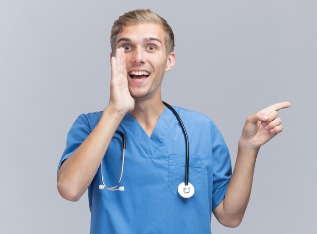 Suspicious young male doctor wearing doctor uniform with stethoscope whispers and points at side isolated on white wall with copy space