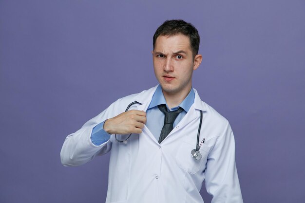 Suspicious young male doctor wearing medical robe and stethoscope around neck looking at camera grabbing his robe isolated on purple background