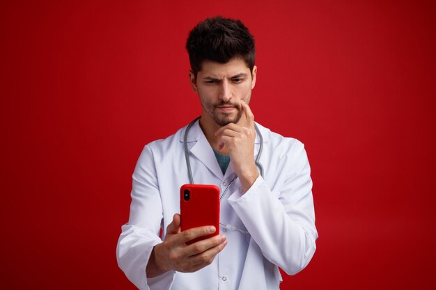 Suspicious young male doctor wearing medical uniform and stethoscope around his neck holding mobile phone touching face talking via video call isolated on red background