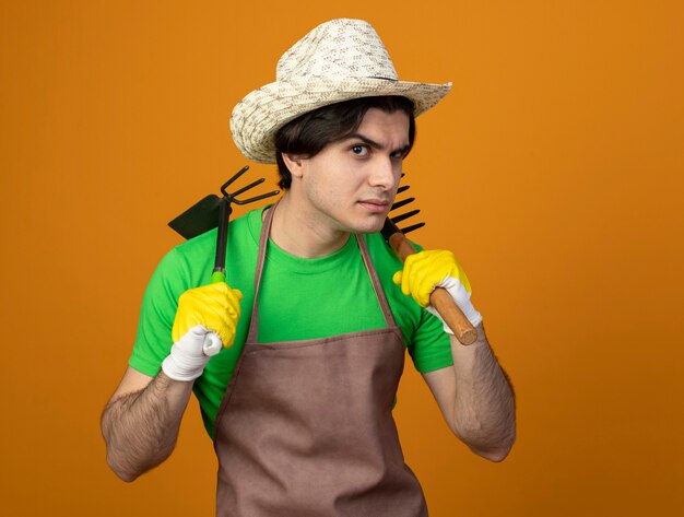 Бесплатное фото Подозрительный молодой мужчина-садовник в униформе в садовой шляпе и перчатках держит грабли с граблями на плече, изолированными на оранжевом