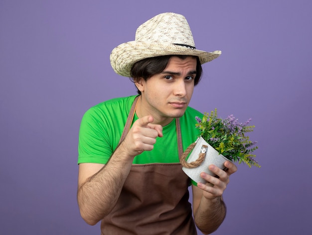 Foto gratuita sospettoso giovane giardiniere maschio in uniforme che indossa cappello da giardinaggio tenendo il fiore in vaso di fiori che ti mostra gesto isolato sulla porpora