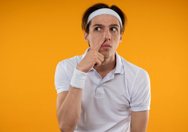 Suspicious young sporty guy looking at side wearing headband and wristband putting finger on nose isolated on orange wall