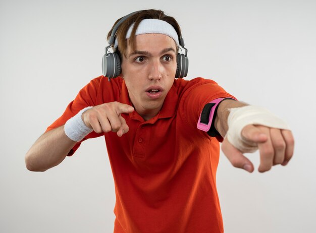 Suspicious young sporty guy wearing headband with wristband and headphones with phone arm band points at side isolated on white wall