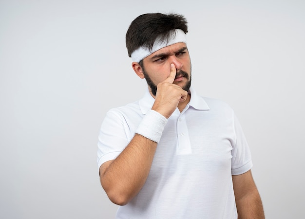 Suspicious young sporty man looking at side wearing headband and wristband putting finger on nose isolated on white wall