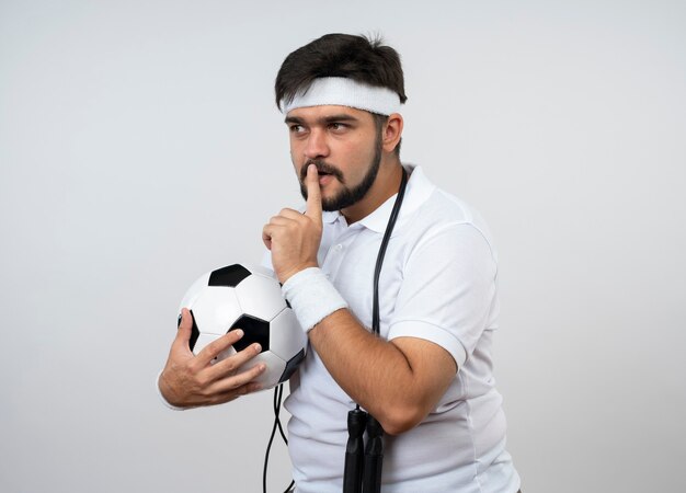 Suspicious young sporty man looking at side wearing headband and wristband with jump rope on shoulder holding ball showing silence gesture isolated on white wall