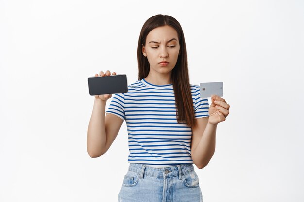 La giovane donna sospettosa che ha dei dubbi, che mostra lo schermo orizzontale dello smartphone, che guarda con incredulità e un sopracciglio mosso alla carta di credito, è preoccupata, in piedi sul muro bianco.