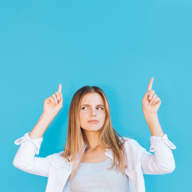 Suspicious young woman pointing her finger upward against blue background