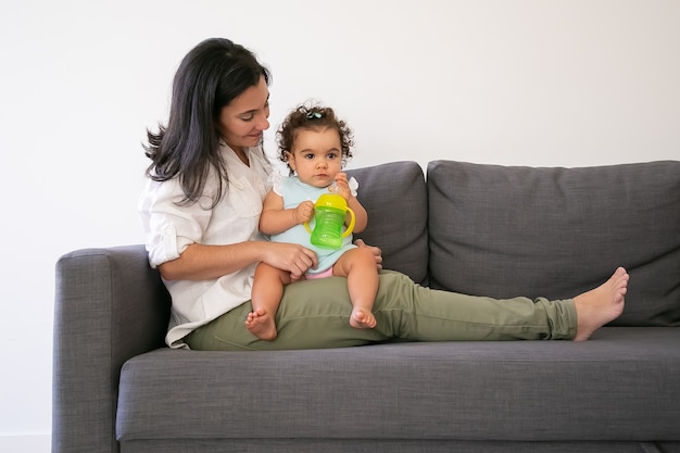 Sweet baby girl sitting on moms lap and drinking water from bottle. Copy space. Parenthood and childhood concept