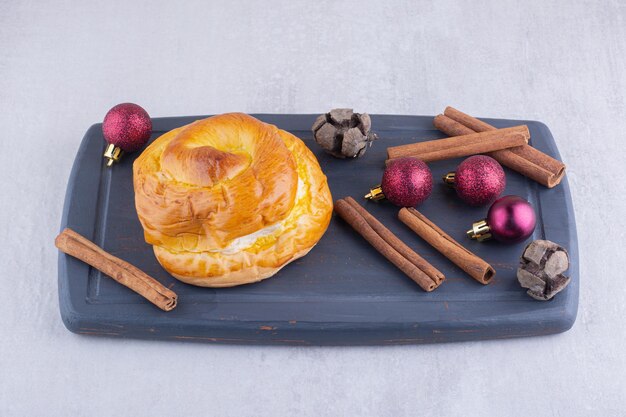 Sweet bun, cinnamon sticks and christmas decorations on a wooden platter on white surface