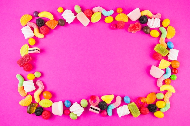 Sweet candies with various shapes forming frame on pink surface