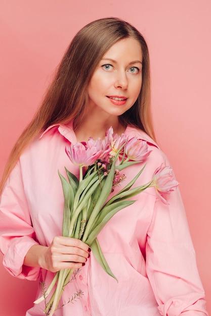 A sweet charming woman with flowers in a pink dress on a pink background smiles happiness and luck