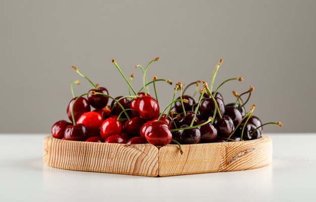 Sweet cherries in a wooden plate on white and grey surface, side view.