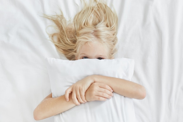 Sweet cute girl with light long hair, hiding behind white pillow, embracing it while lying in bed, having fun in morning