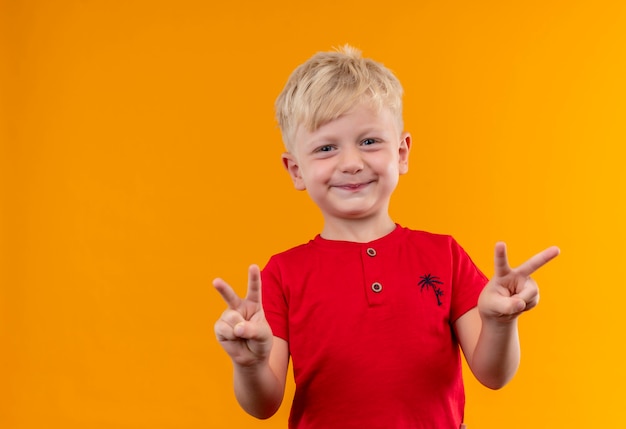 A sweet cute little boy with blonde hair and blue eyes wearing red t-shirt showing two fingers gesture