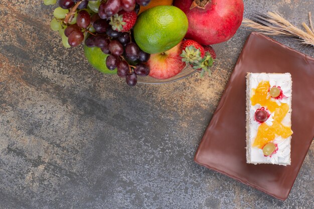 Sweet different fruits on glass plate with piece of cake on dark plate