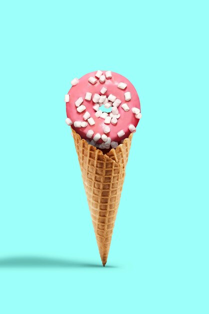 Sweet donut glazed by pink icing sprinkled with marshmallow in a wafer cone against turquoise background. Concept of food, treats and unhealthy nutrition. Close up, copy space