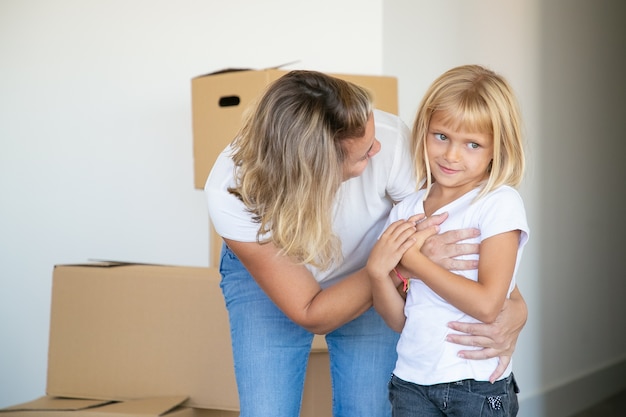 Sweet fair haired girl and her mom moving into new flat, standing near stack of boxes and hugging