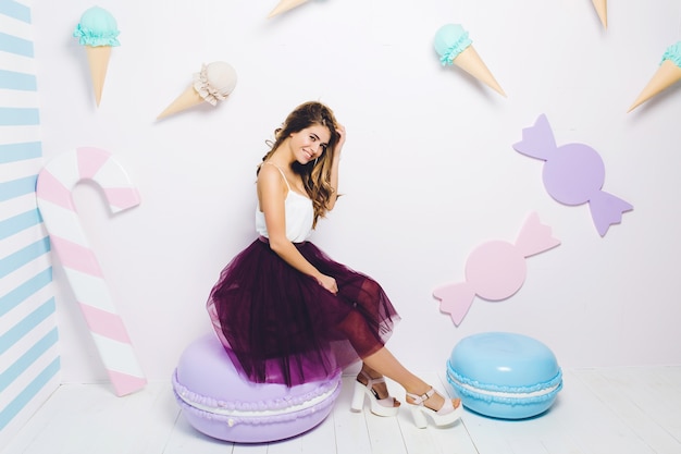 Sweet happy summer time of attractive fashionable young woman in tulle skirt sitting on huge macaron. Pastel colors, sweets, delicious, enjoying, happiness, smiling, relaxing.