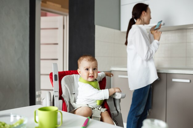 Sweet little baby in green bib sitting in baby chair getting scared seeing bird outside the window while mom turns aside to check chat rooms on smartphone.