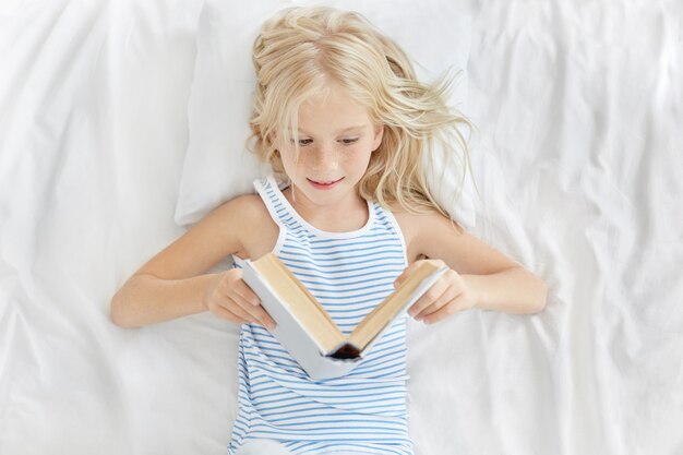 Милая блондинка 7-летней европейской внешности отдыхает в белой кровати, с интересом смотрит в открытую книгу и читает сказку
