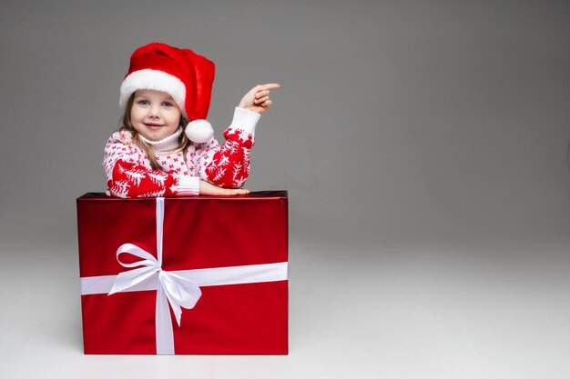 Sweet little girl in patterned winter sweater and Santa hat indicating at blank space leaning on wrapped Christmas present with white bow.