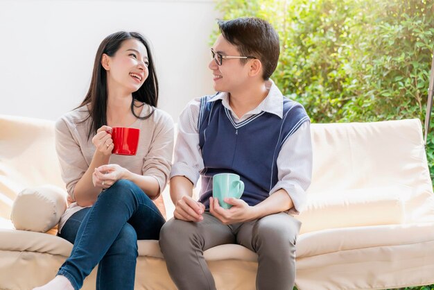 달콤하고 사랑스러운 아시아 부부의 남편과 아내는 뜨거운 음료를 마시며 행복과 평화로운 가정 내부 배경과 함께 이야기를 나눕니다.
