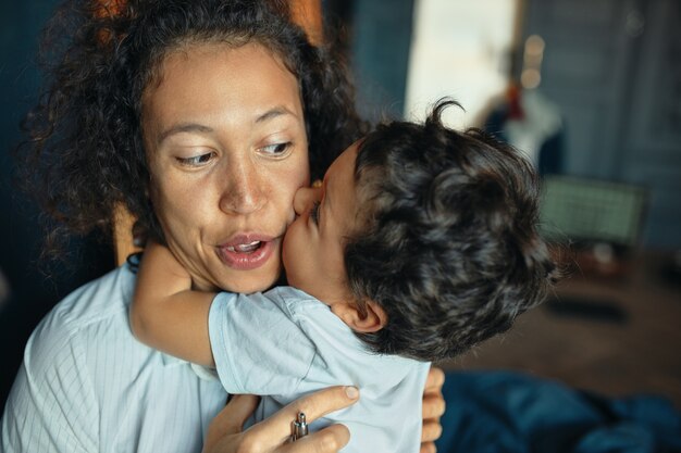 Сладкий портрет милого нежного маленького мальчика смешанной расы, целующего молодую свою возбужденную маму в щеку, обнимая ее за шею.