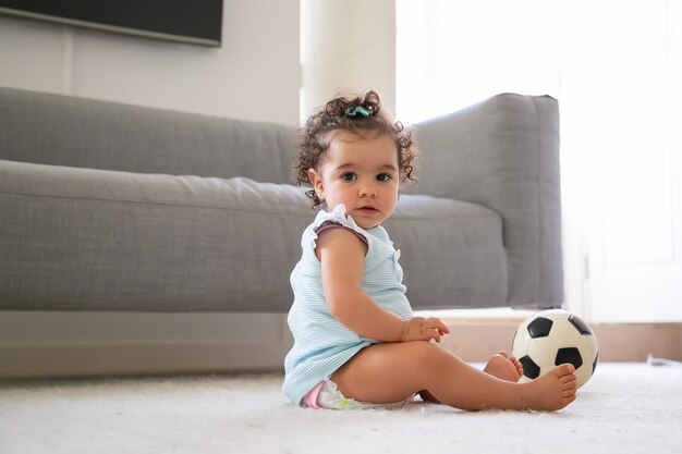 Милая серьезная черноволосая девочка в бледно-голубой одежде сидит на полу с футбольным мячом, a. Вид сбоку. Ребенок дома и концепция детства