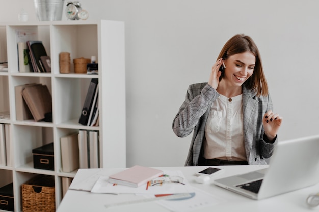 Sweet short-haired business lady laughs while listening to music on headphones. portrait of woman in office clothes sitting in workplace.