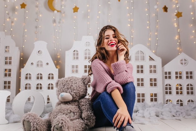 Sweet, smiling, young girl in cozy is sitting on floor with teddy bear