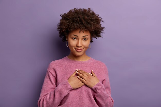 sweet thank you. charming touched sincere afro american woman appreciates surprise or adorable gift, keeps palms pressed to heart, being very grateful to you, poses against purple wall
