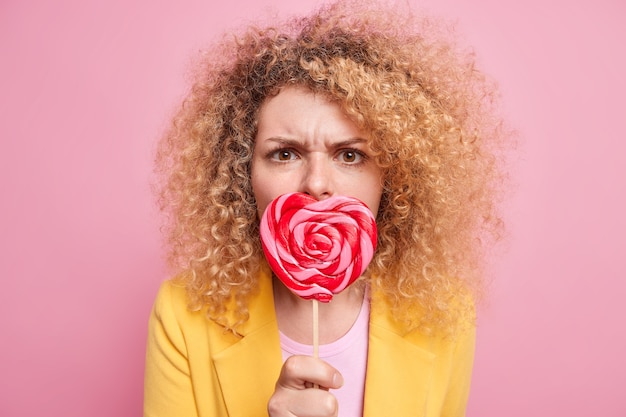 Sweet tooth concept. Serious curly haired young European woman covers mouth covers mouth with delicious lollipop has worried expression focused displeased poses against pink wall.