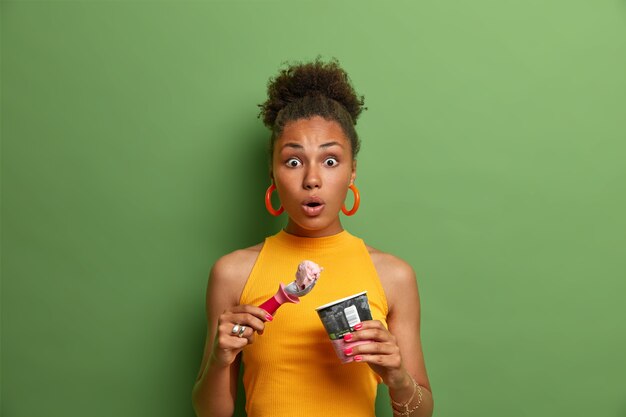 Sweet tooth and temptation concept. Speechless surprised Afro American woman eats tasty cold dessert, enjoys ice cream with strawberry flavor, dressed in yellow summer clothes, green wall