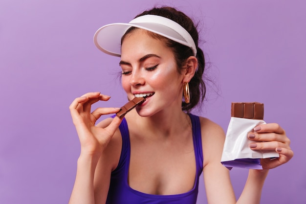 Foto gratuita dolce donna in berretto e top viola morde con piacere cioccolato al latte