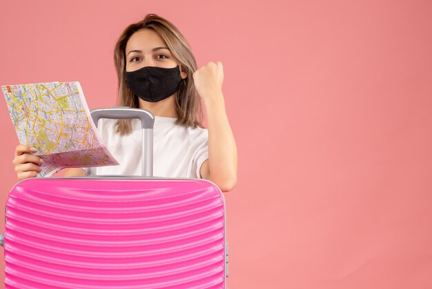 sweet young woman with black mask holding map behind pink suitcase