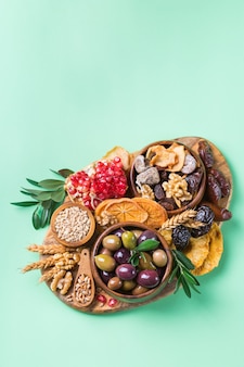 Symbols of judaic holiday tu bishvat, rosh hashana new year of the trees. mix of dried fruits, date, fig, grape, barley, wheat, olive, pomegranate. copy space flat lay green background